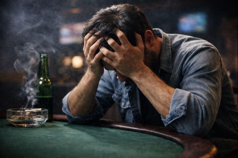 A man sits alone at a dim bar with his head in his hands, looking distressed, with a beer bottle and an ashtray on the table.