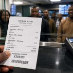 Football betting slip at a betting shop counter, showing a close-up payout receipt behind glass with a card terminal and blurred customers in the background.
